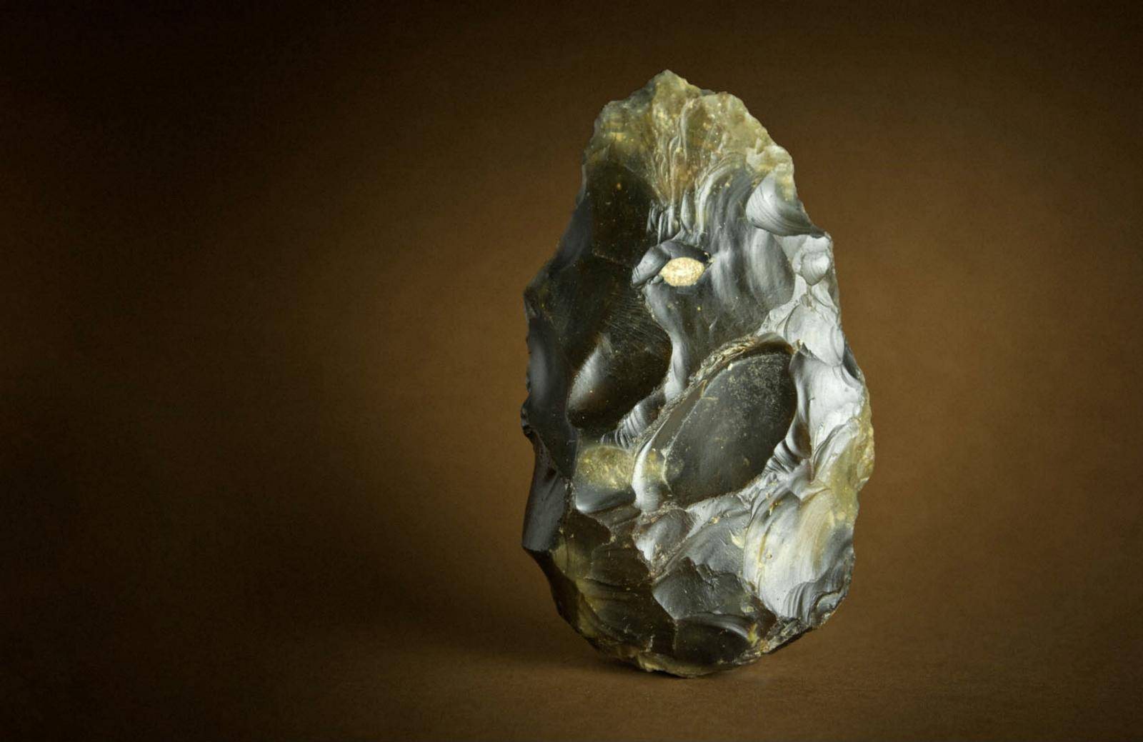 Studio photograph of a knapped flint hand axe against a warm brown background